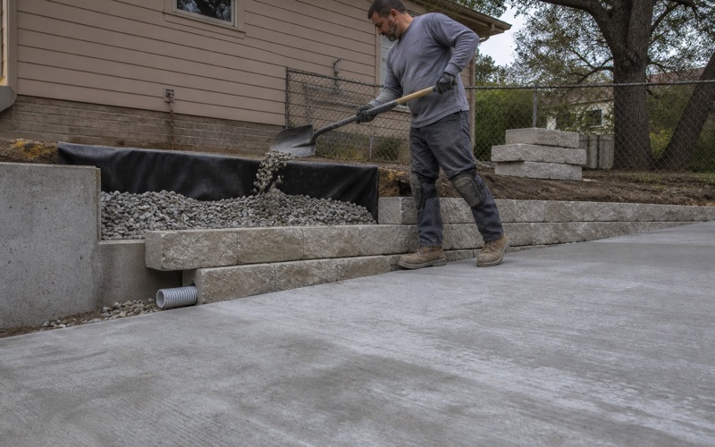 Drainage stone being shoveled in behind a part-built block retaining wall in Temple, filter fabric up the back and an outlet pipe at the low end.
